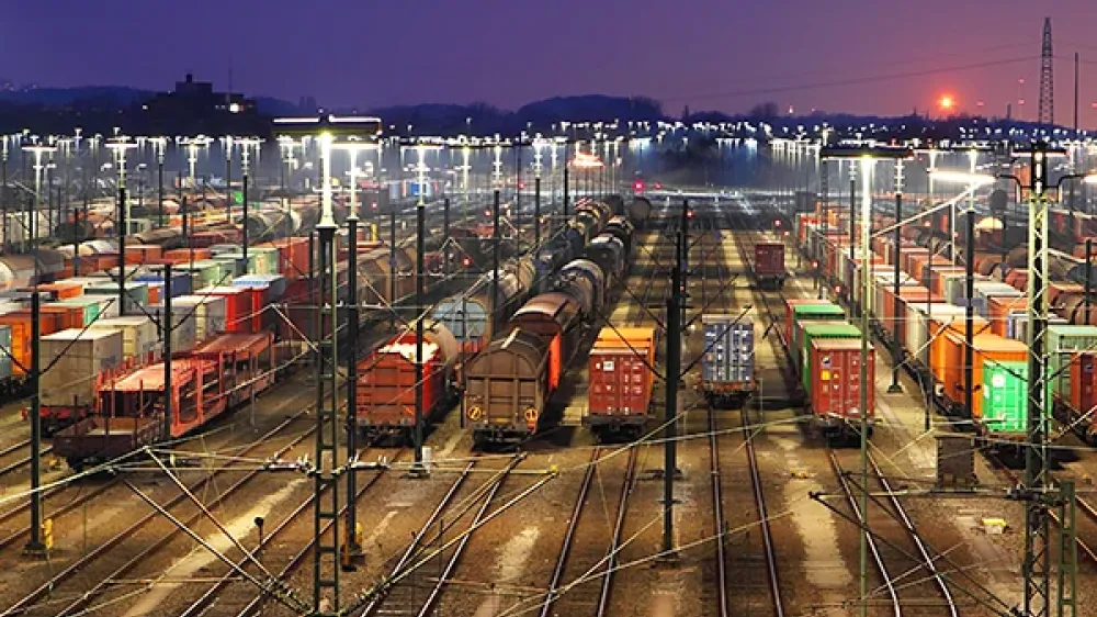The railway station has many tracks, points and power lines in front of the silhouette of a city.
