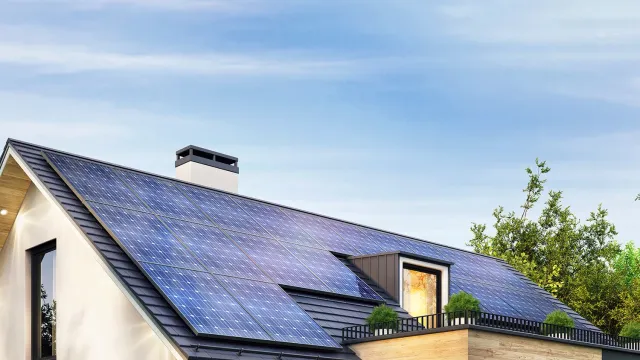 Modern single-family house with rooftop solar panels, large windows, and surrounding trees under a clear blue sky.