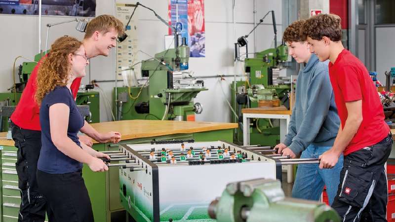 Four young people, two on each side, play foosball in an industrial workshop. They are smiling and focused, with green machinery in the background.