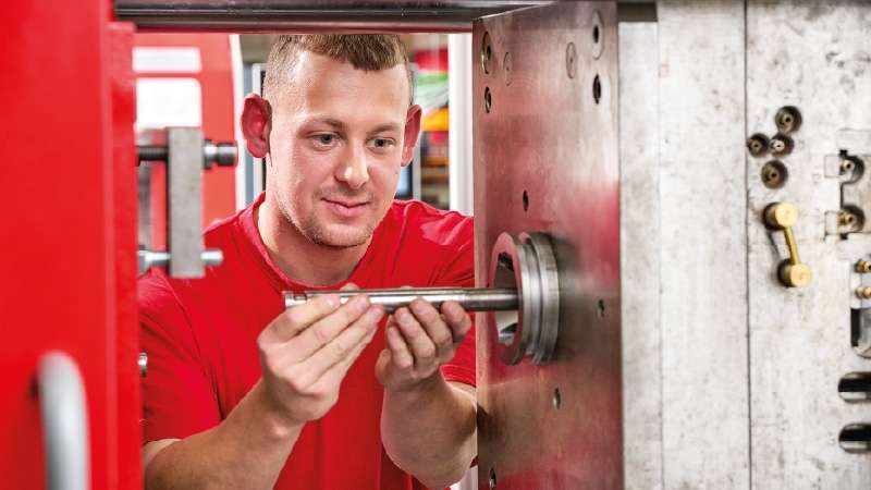A man in a red shirt inspects or assembles a metal component on industrial machinery in a workshop setting.