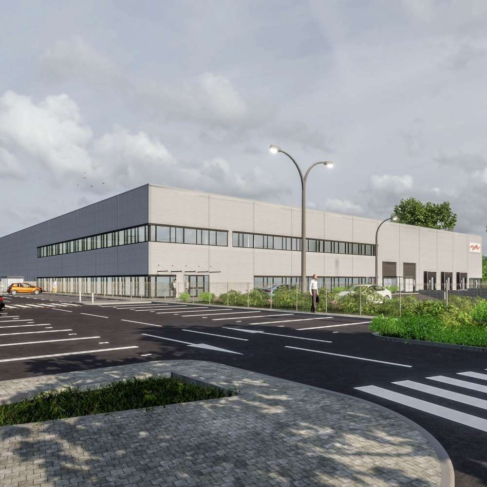 A modern, two-story industrial building with large windows, a parking lot in front, streetlights, and greenery, under a cloudy sky.