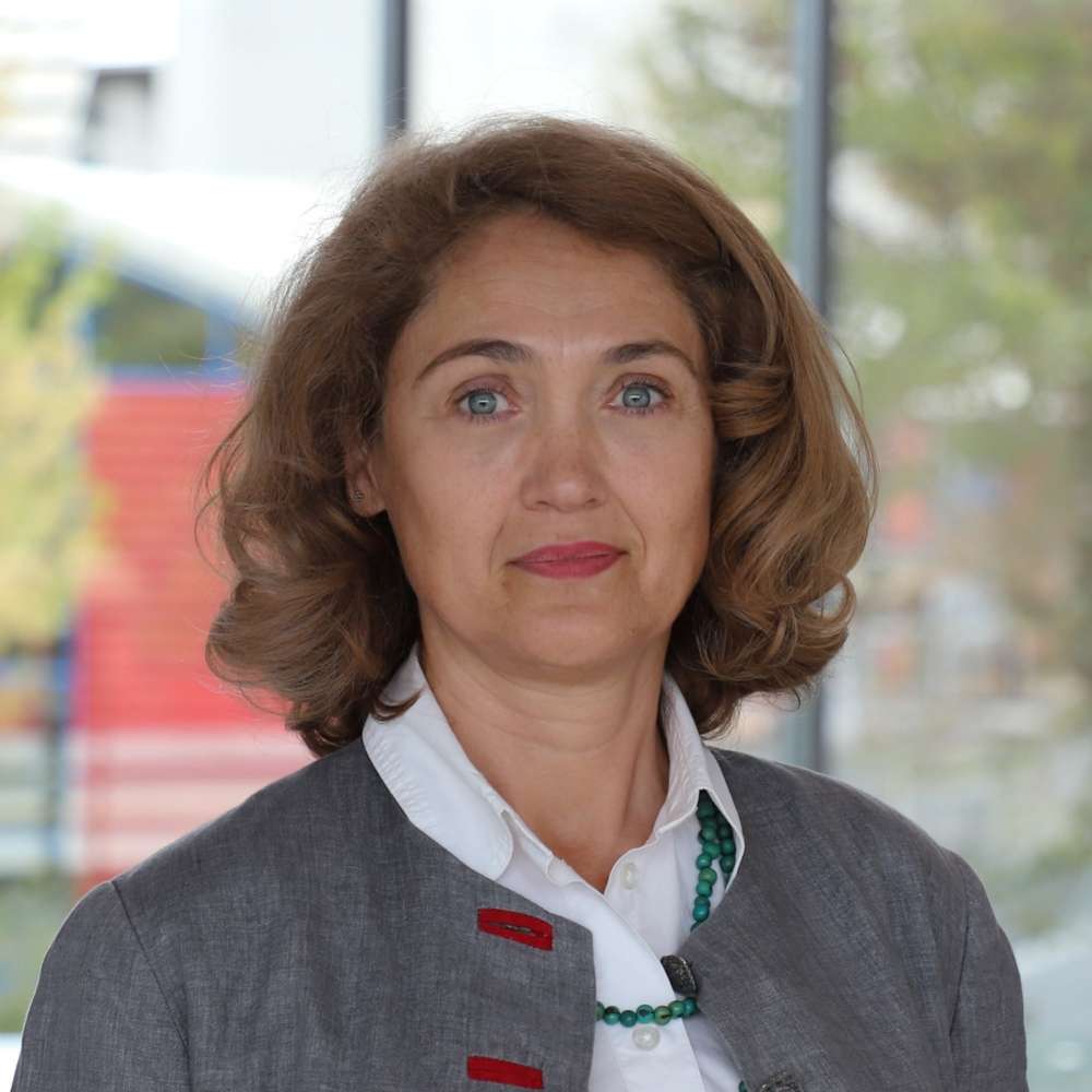 A captivating image displays a woman in a sleek gray blazer, her wavy brown hair elegantly complementing the professional setting. The green and red beaded necklace adds a pop of color, while the blurred background of a building and trees hints at a sophisticated indoor environment.