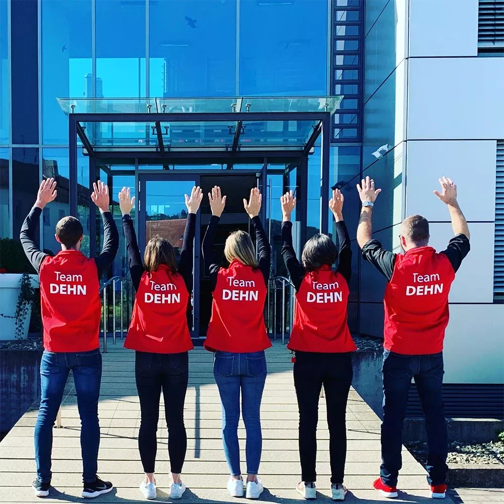 Five employees wearing red “Team DEHN” vests stand in front of a modern office building with their arms raised.