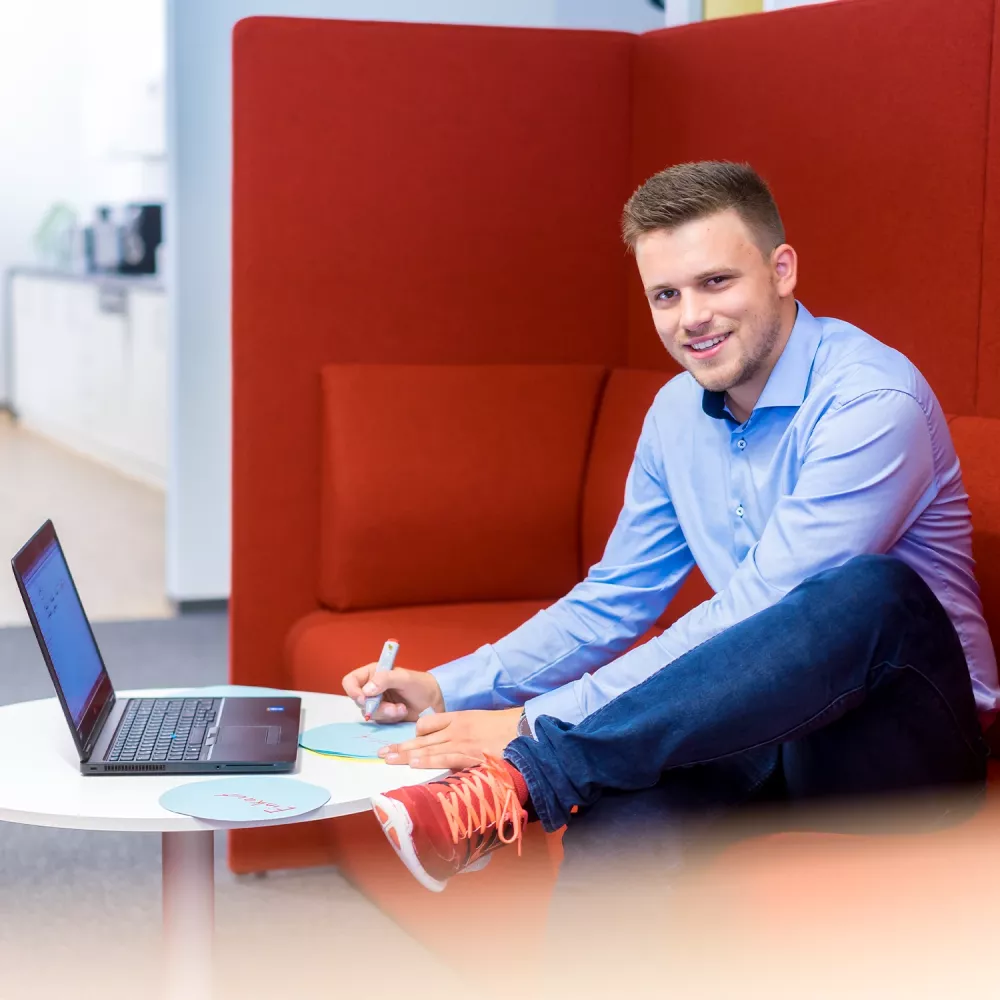 Young man sitting on an orange lounge sofa, working with a laptop and writing notes at a small table in a modern office.