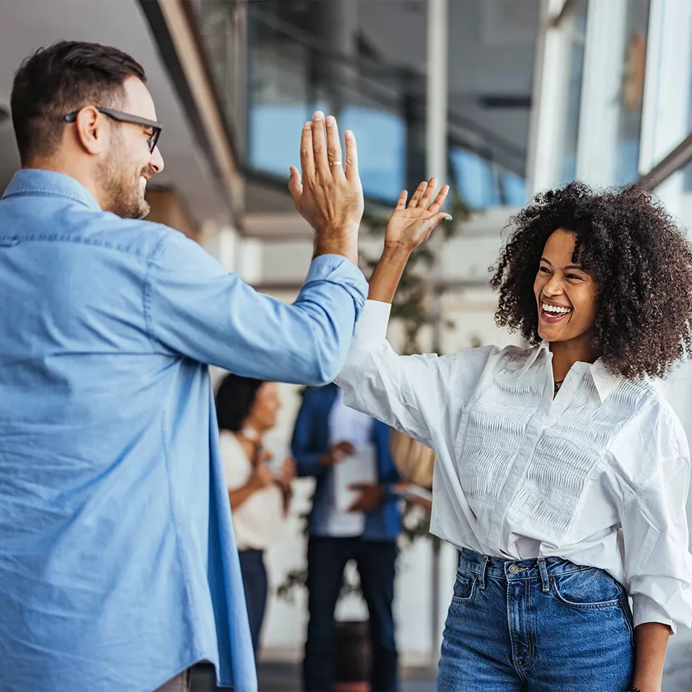 Two colleagues smiling and giving a high-five in a modern office environment, with coworkers blurred in the background.