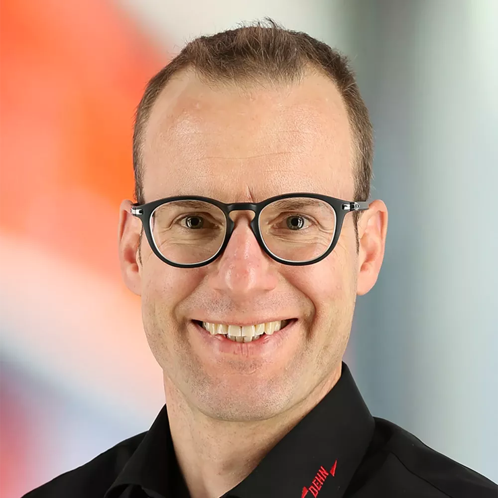 Portrait of a smiling man wearing round glasses and a black shirt, photographed against a softly blurred background.