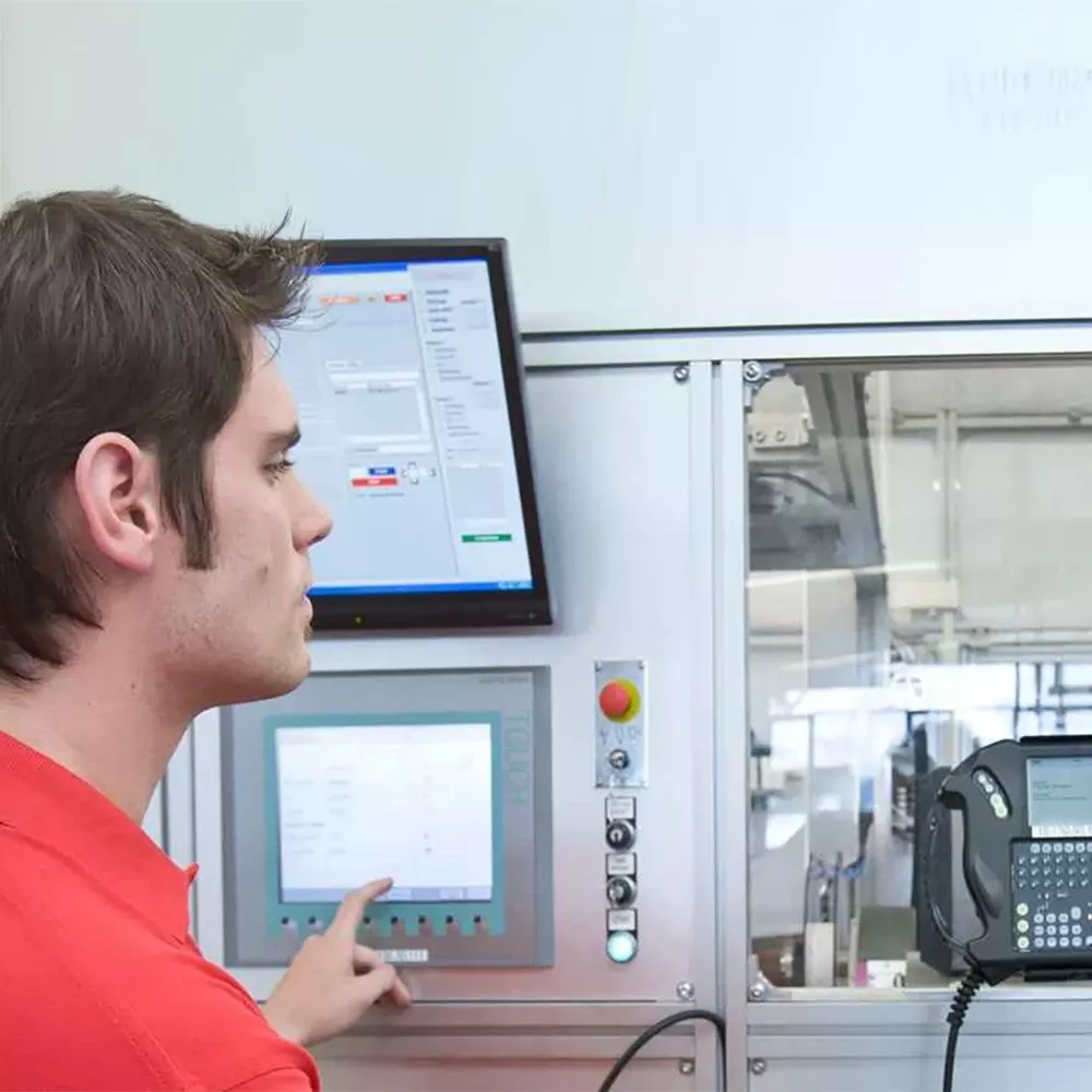 Man operating an industrial control panel with a touchscreen interface, standing next to enclosed machinery and a mounted display screen.