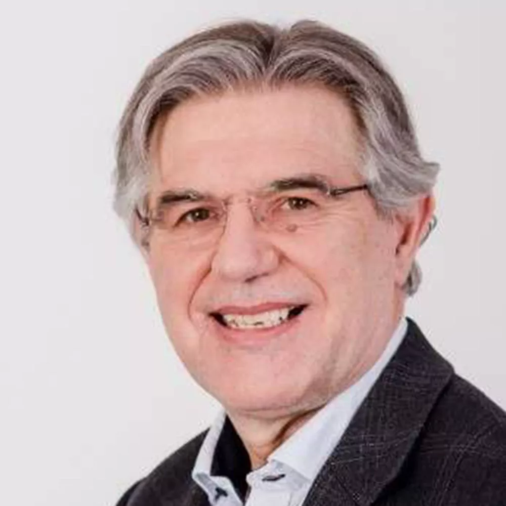 Portrait of a gray-haired man wearing glasses and a dark jacket over a light shirt, smiling against a white background.