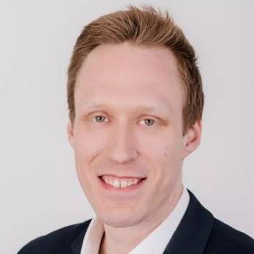 Portrait of a man with short light brown hair wearing a dark jacket and light shirt, smiling against a white background.
