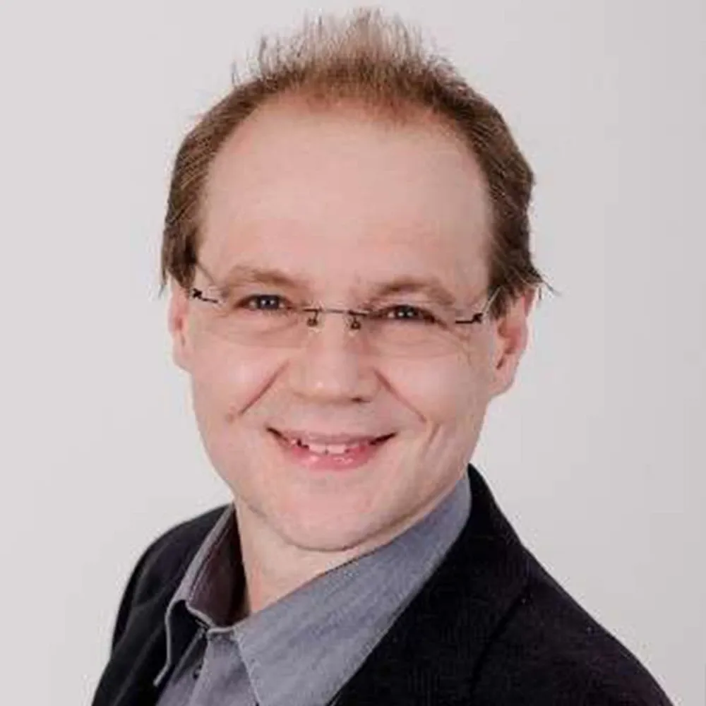 Portrait of a man wearing glasses, a dark jacket, and a gray shirt, smiling against a white background.