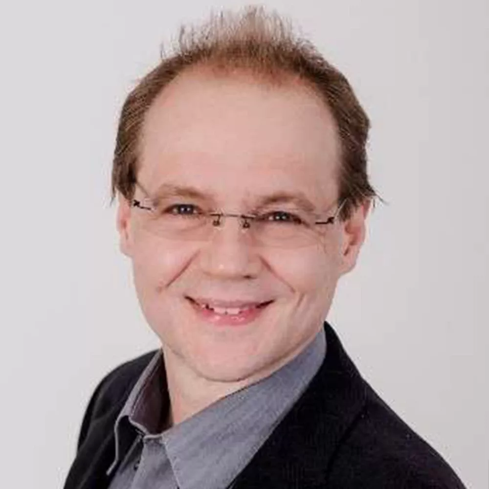 Portrait of a man wearing glasses, a dark jacket, and a gray shirt, smiling against a white background.