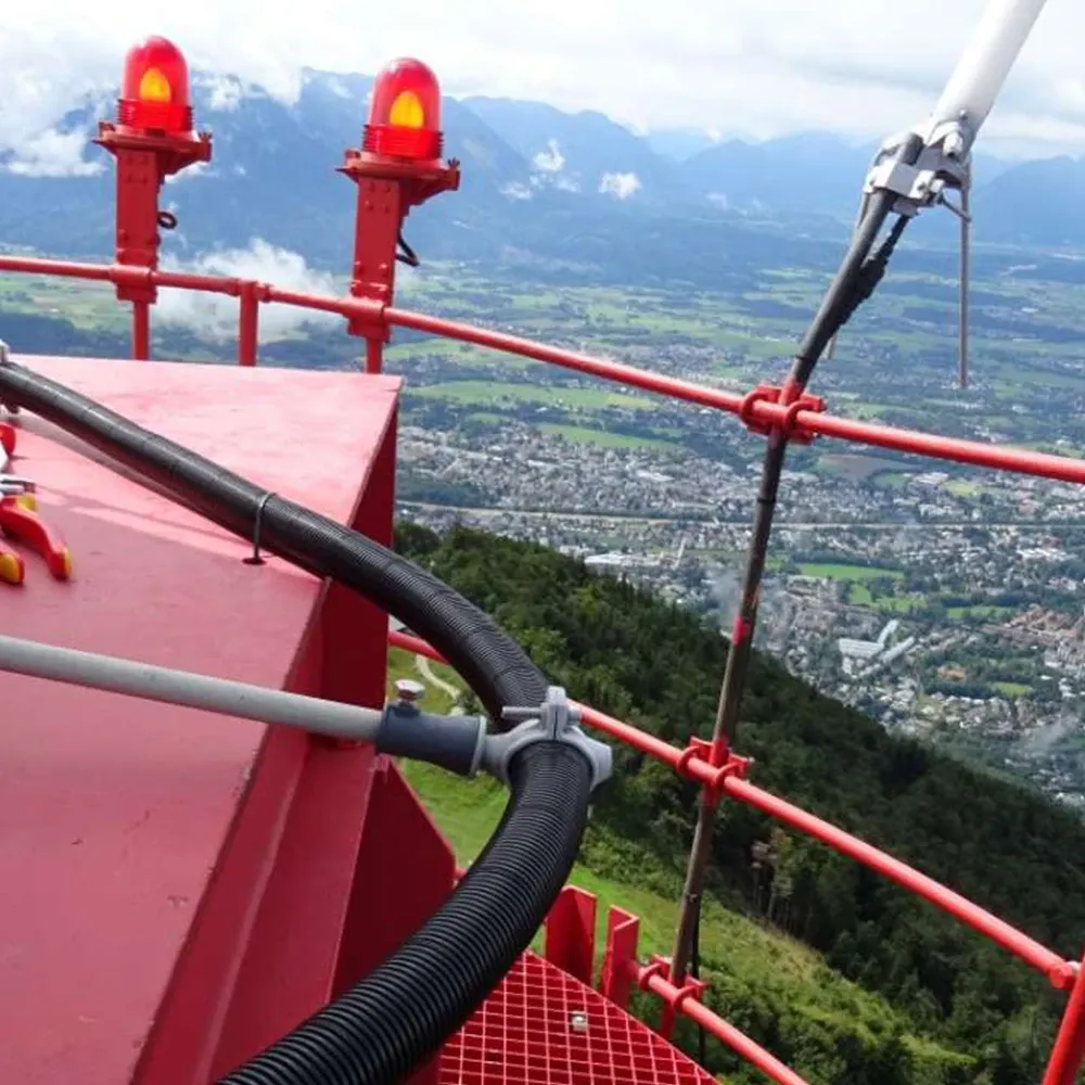 Red metal railing with warning lights and cables on a tower platform, overlooking a valley with buildings, fields, and mountains.