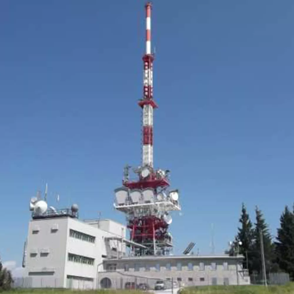 Tall red-and-white transmission tower with multiple antenna dishes beside a building, set against a clear blue sky.