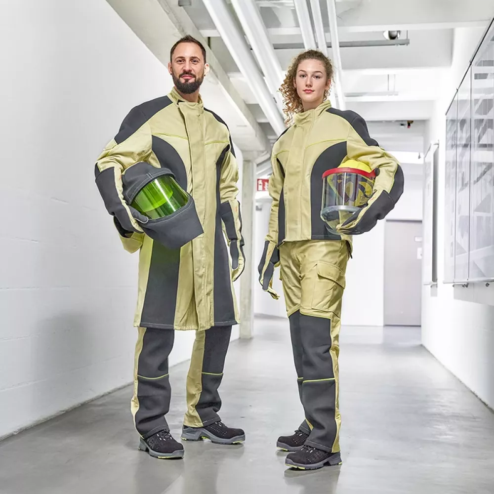 Two electricians in protective workwear stand in an industrial corridor, holding helmets with visors as part of modern personal protective equipment.
