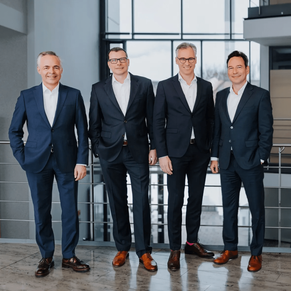 Four men in suits standing side by side in a modern interior with large windows in the background.
