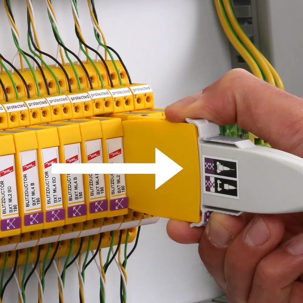 Hand removing a yellow surge protection module from a control cabinet while holding a white test adapter; white arrow indicates direction.