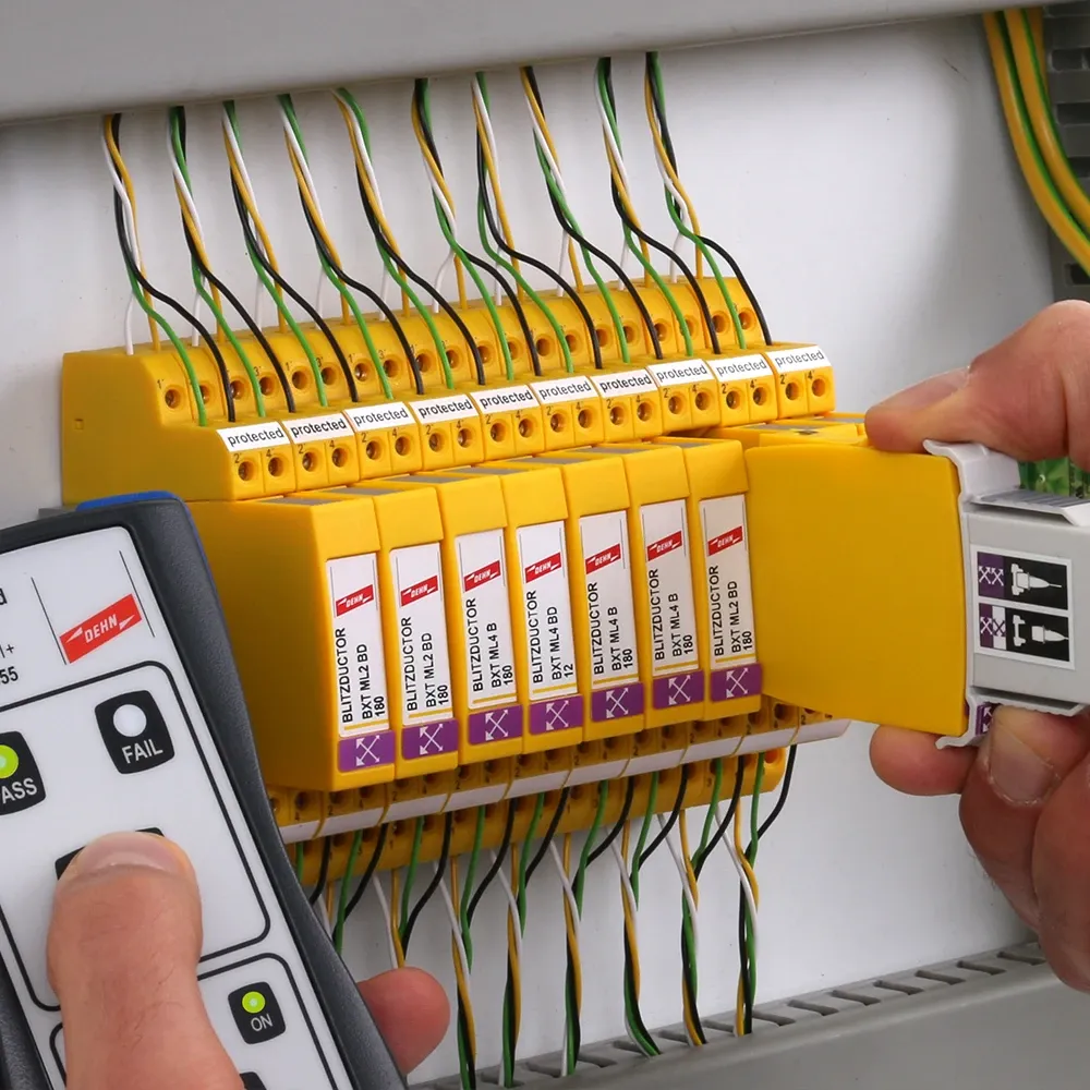 Hand holding a measuring device next to a row of yellow surge protection modules while another hand holds a removed module with test adapter.