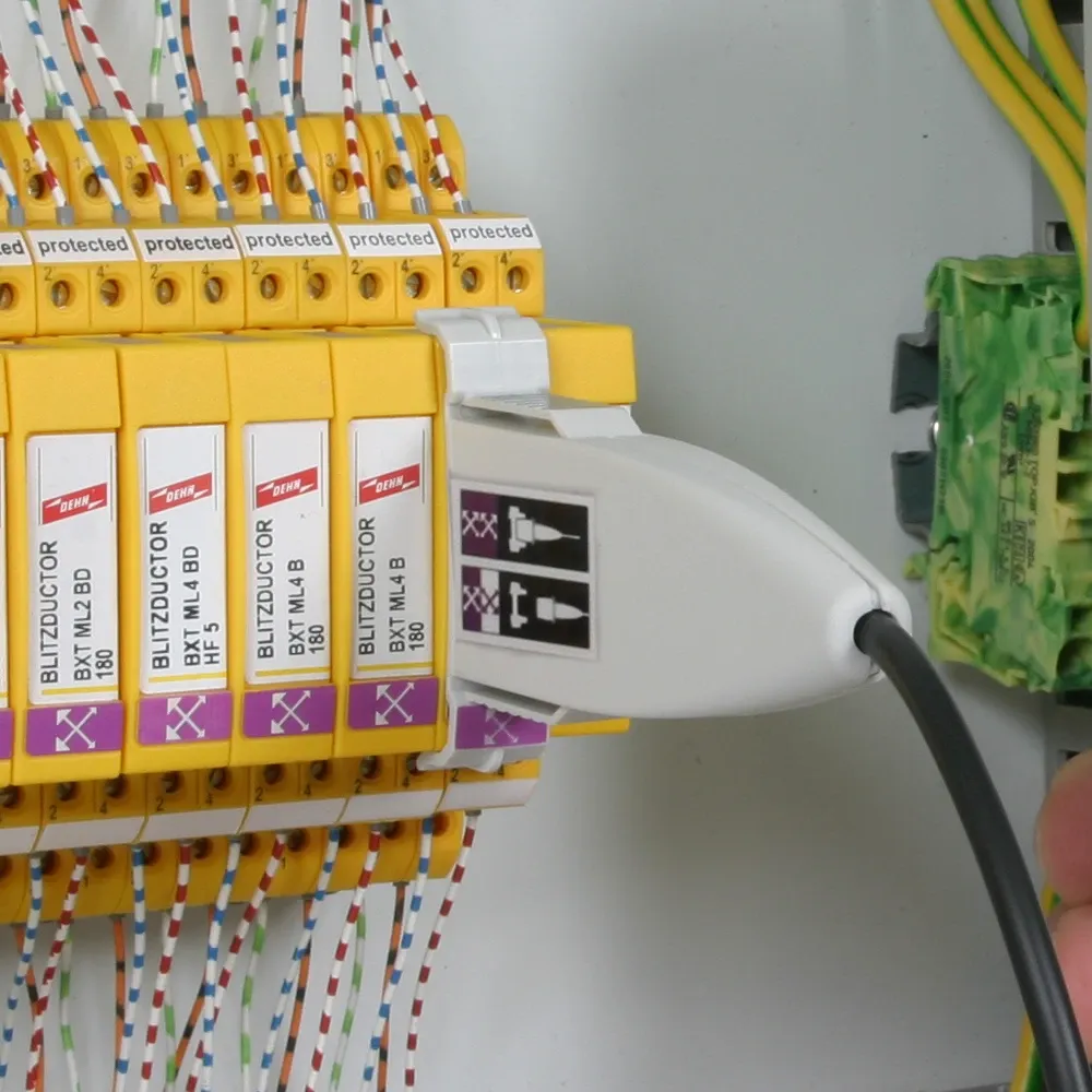 White test adapter connected to a yellow surge protection module inside a control cabinet, with cable attached and wires visible.