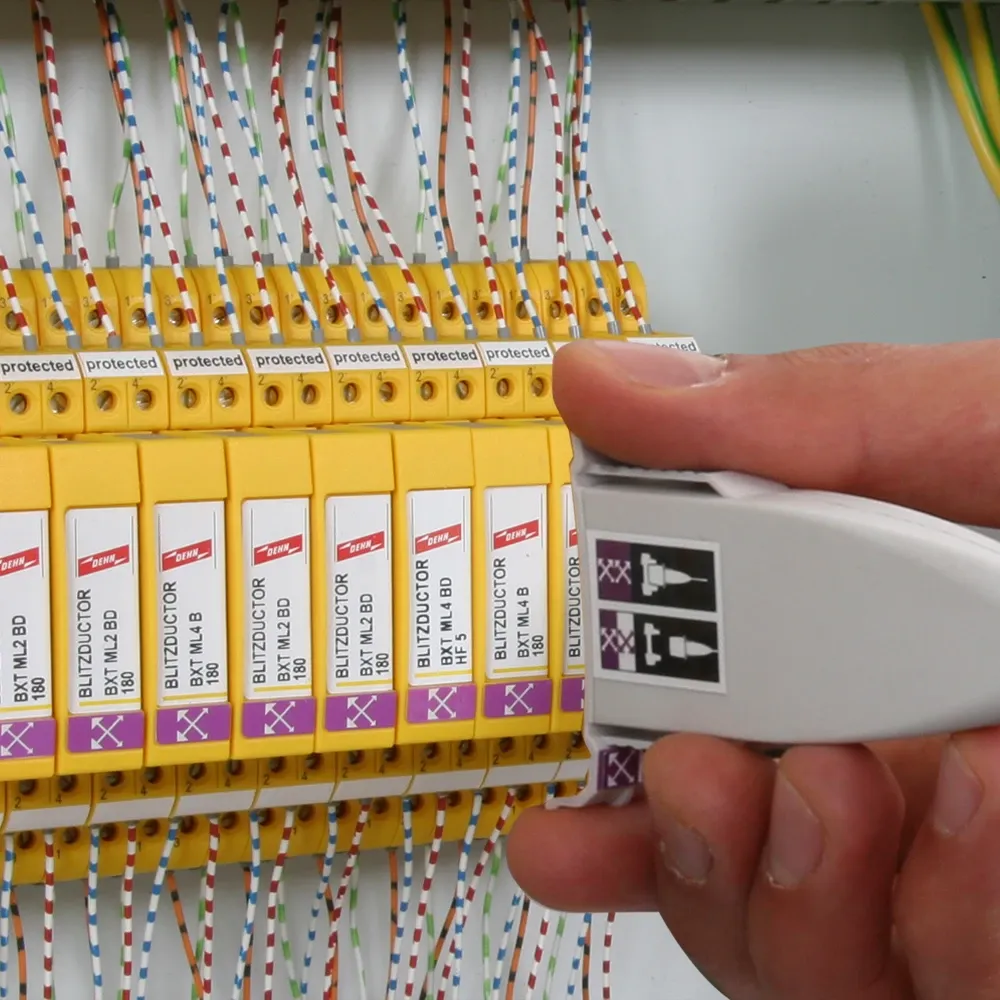 Hand holding a white test adapter next to yellow surge protection modules inside a control cabinet with connected wires.