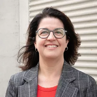 Portrait of a smiling woman wearing glasses, a gray blazer, and a red top, standing in front of a light-colored wall.