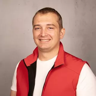 Smiling man wearing a red sleeveless vest over a white T-shirt, standing in front of a light gray background.