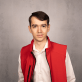 A striking image displays a young man in a vibrant red vest, his short, dark hair and friendly gaze standing out against a clean gray wall. The well-composed shot highlights the contrast of his white shirt and black zipper, making for an engaging and eye-catching portrait.