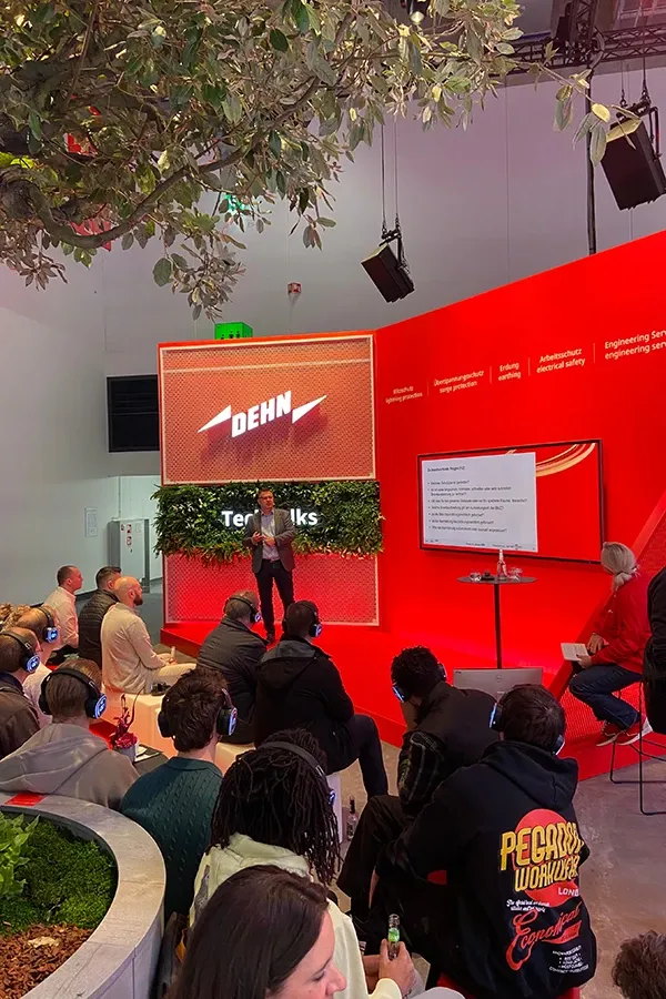 A speaker presents at a DEHN event in front of a seated audience, with a large screen displaying slides and red walls featuring company branding.