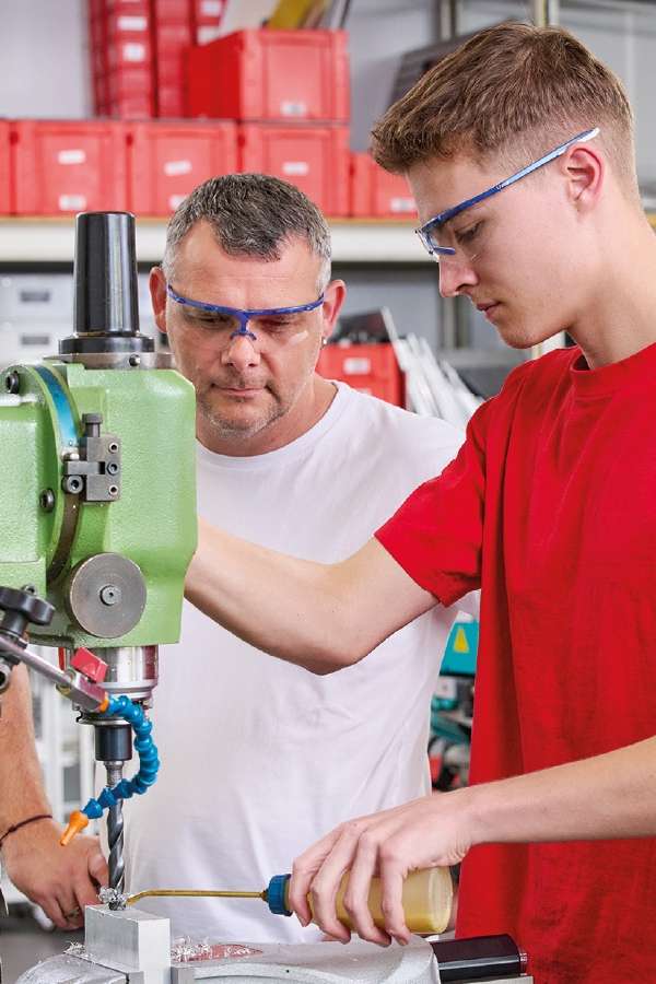 Two men wearing safety glasses work with a green drill press in a workshop. One operates the machine, the other supervises, focusing intently.