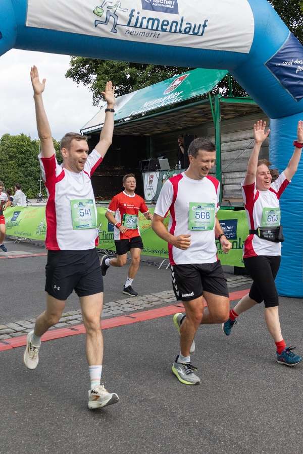 Three runners in white and red shirts celebrate with arms raised as they cross the finish line under a blue arch at a vibrant outdoor race event.
