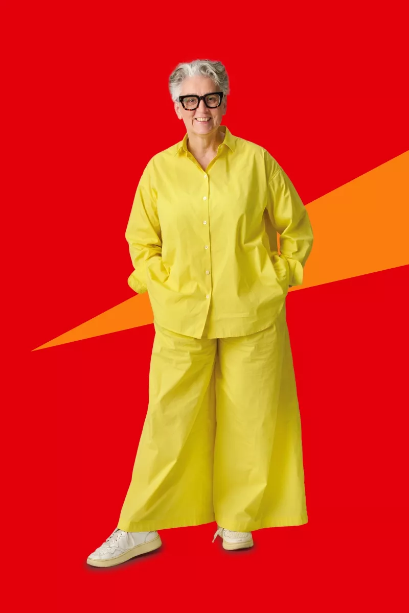 Person standing with hands in pockets wearing a loose yellow shirt and wide yellow pants against a red background with an orange diagonal shape