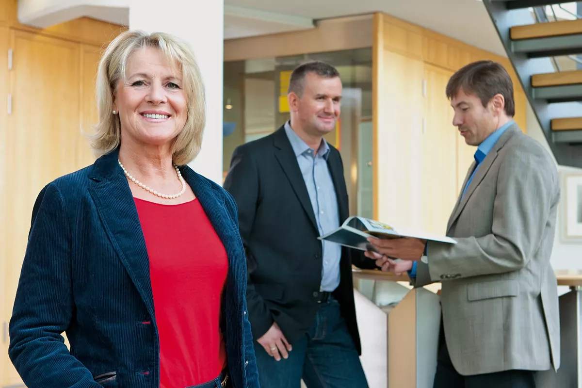 Smiling businesswoman standing in the foreground of a modern office, with two male colleagues in the background discussing documents near a staircase.