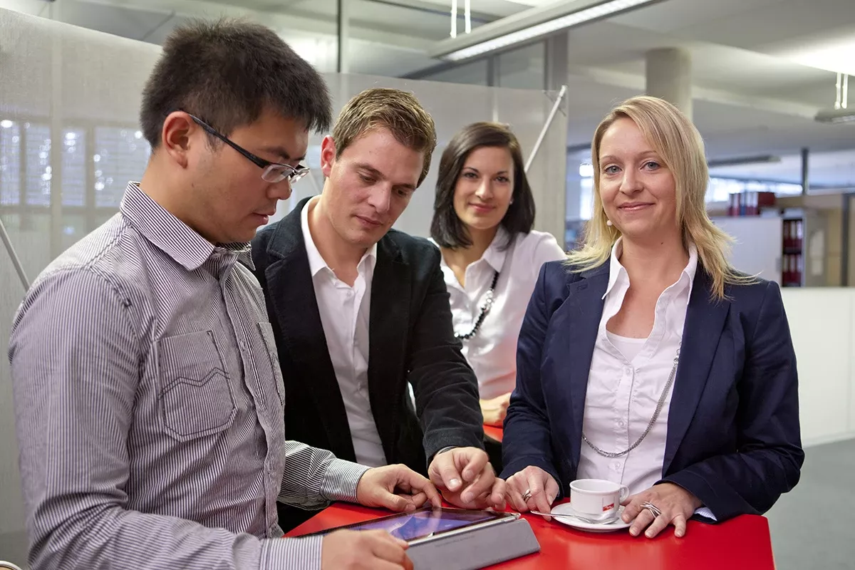 Four colleagues gathered around a tablet at a red table in a modern office, discussing content together, with a coffee cup visible in the foreground.