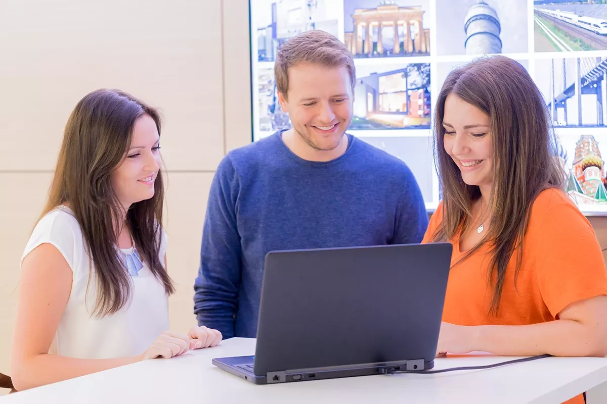 Three students standing together at a table, smiling and looking at a laptop screen during a collaborative study session.