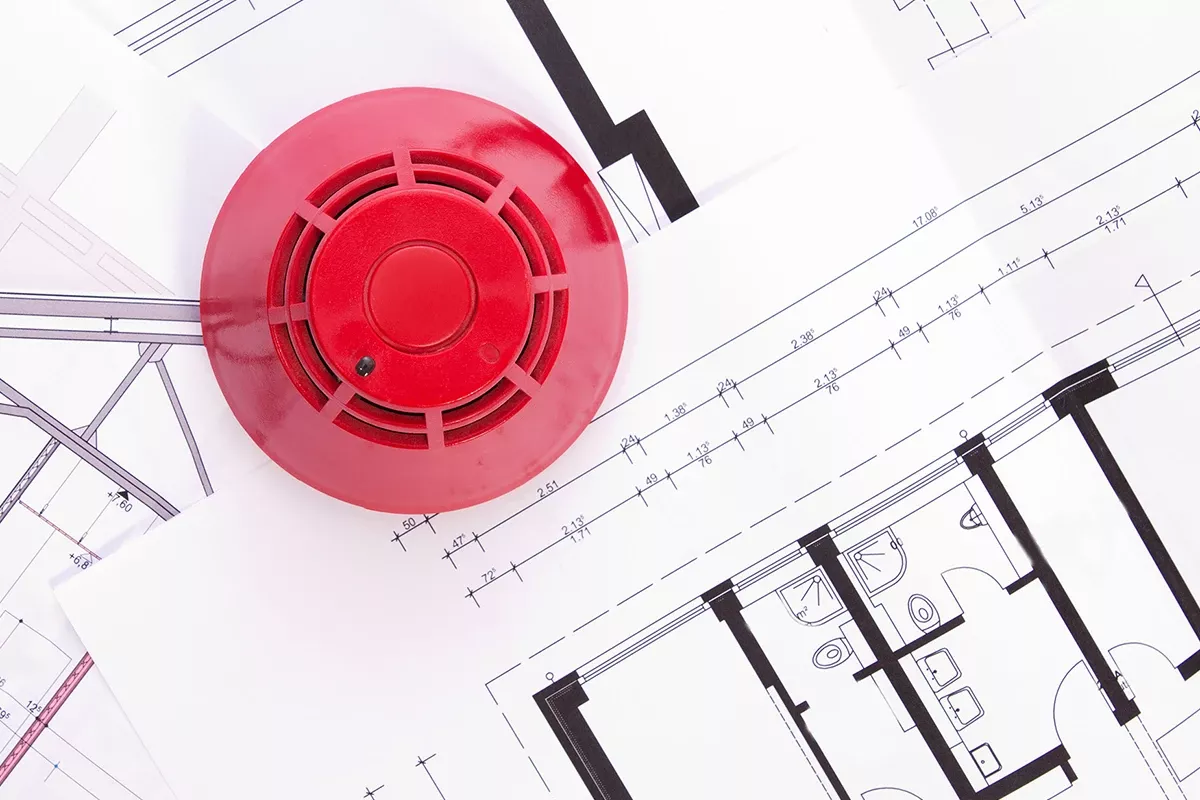 Red fire alarm device placed on architectural floor plans with technical drawings and measurements visible.